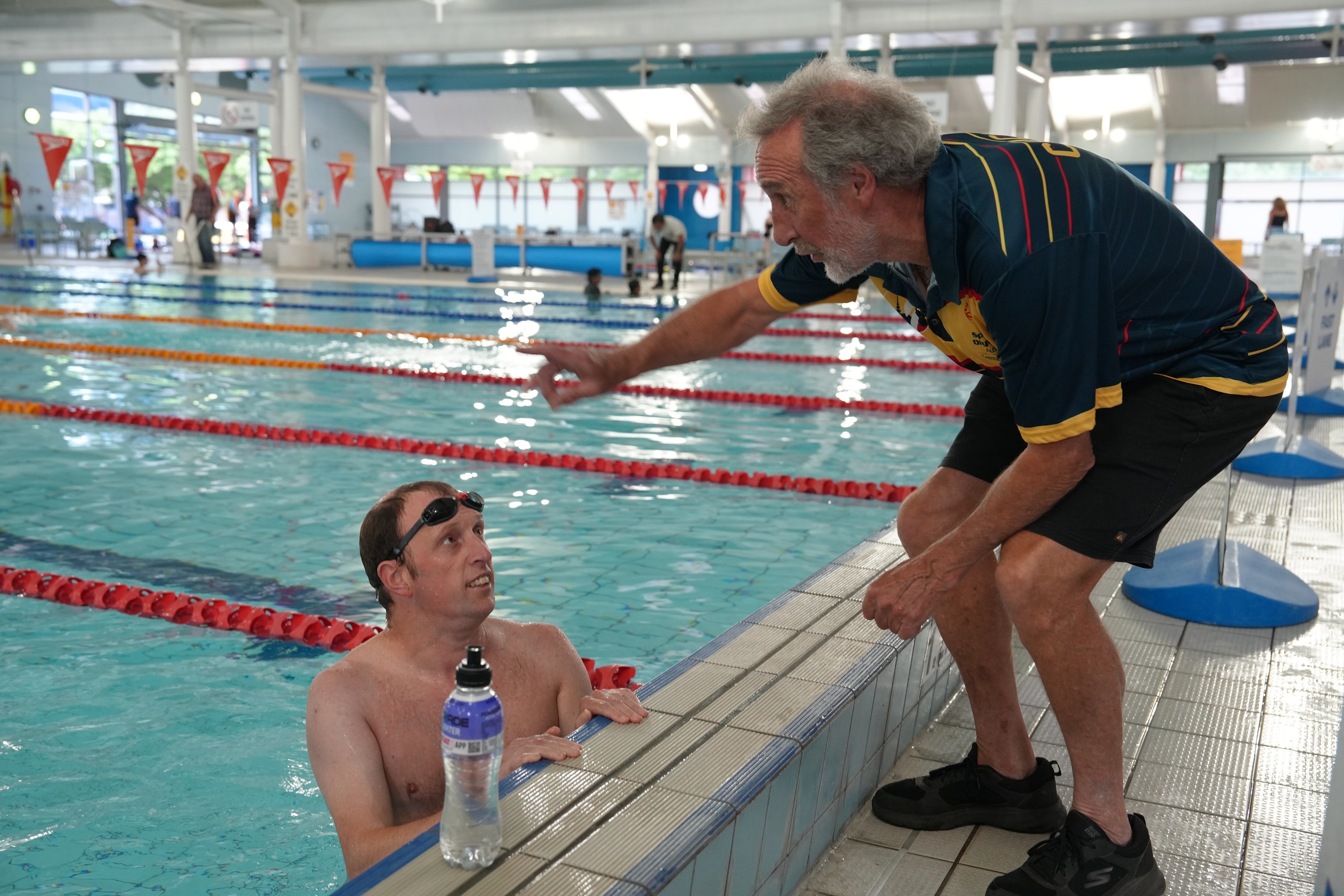 A man in an indoor lane pool looks out of the water at another man as they speak to one another.