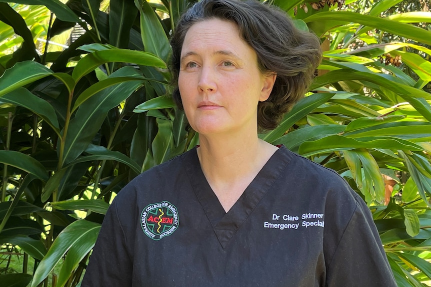 Dr Skinner looks out of frame, standing in front of a large lush green bush. She is wearing navy ED scrubs.