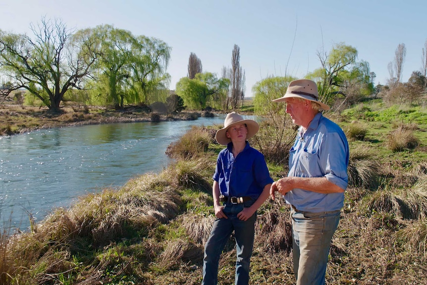 A teenager and his grandfather stand next to a flowing river. Both wear wide-brim hats