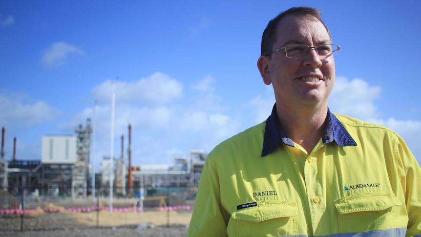 A man in a high-vis shirt, with glasses. An industrial plant is behind him