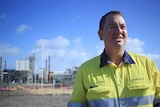 A man in a high-vis shirt, with glasses. An industrial plant is behind him