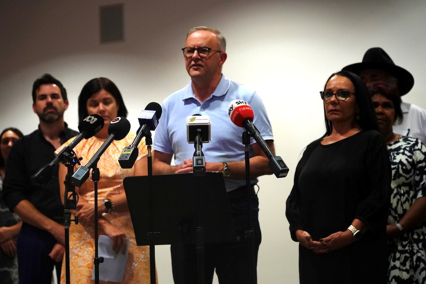 Anthony Albanese stands in front of microphones as other people watch from either side. 