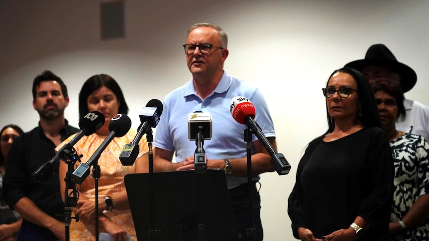 Anthony Albanese stands in front of microphones as other people watch from either side. 