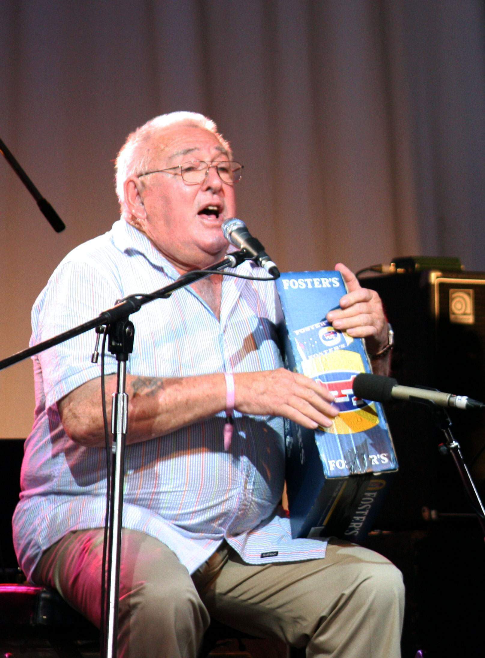 Ted Egan está sentado frente al micrófono y sostiene un cartón de cerveza en la mano.