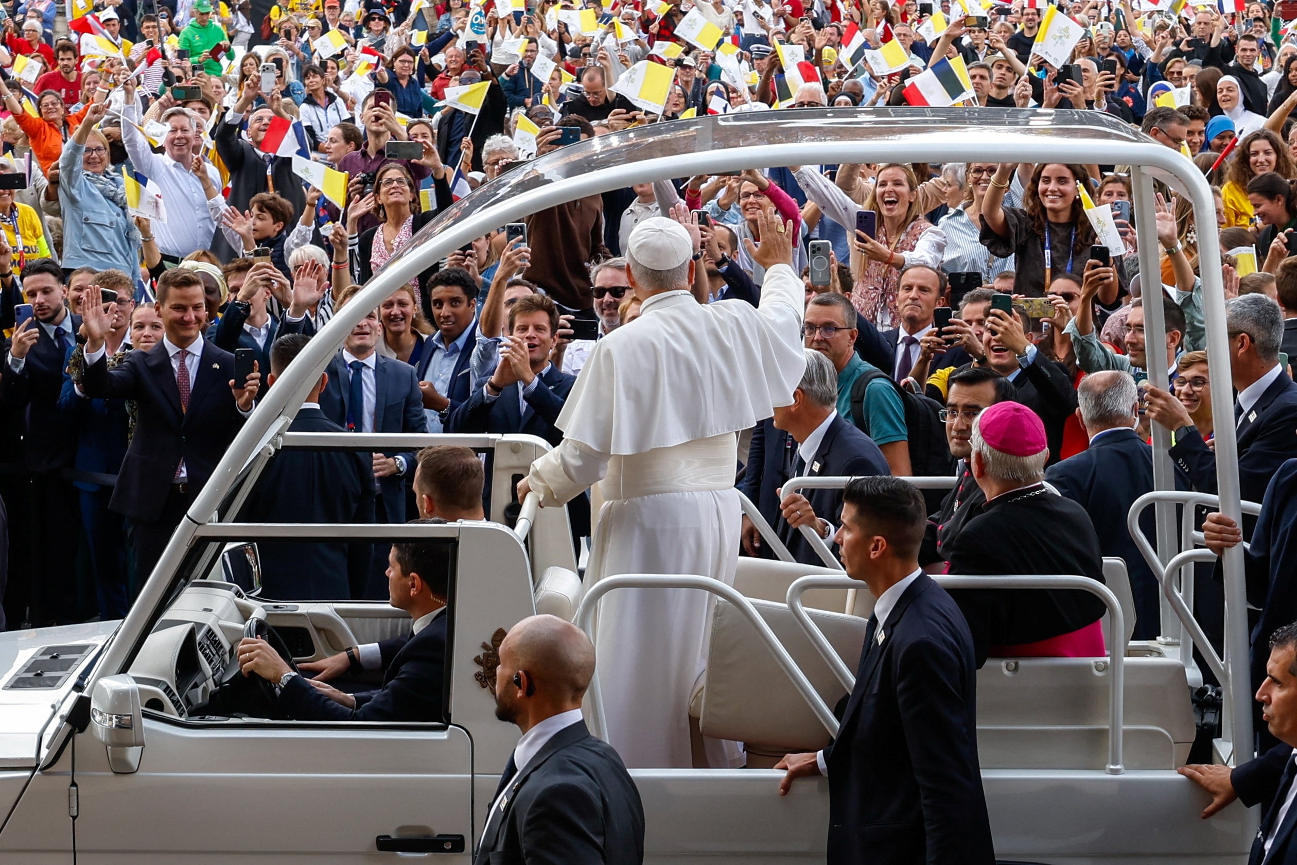 The pope stands back to camera and waves at a large crowd waving flags as he drives by in his Popemobile.