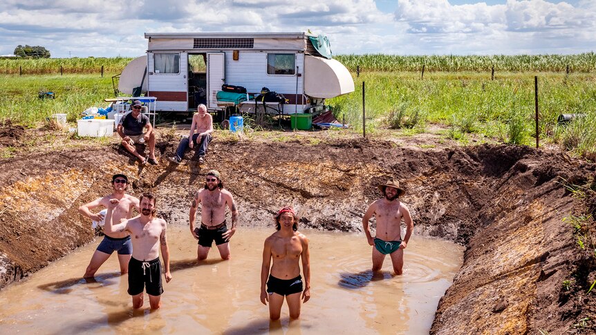 A group of shirtless men stand in a muddy hole by a caravan.