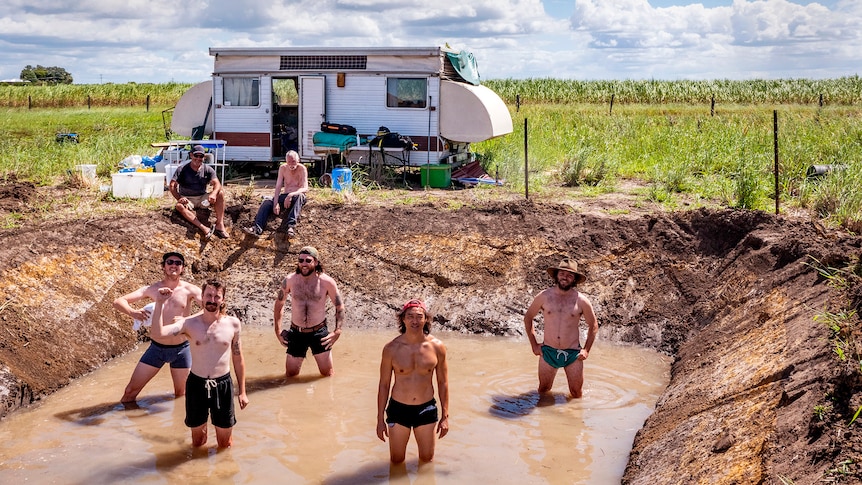 A group of shirtless men stand in a muddy hole by a caravan.