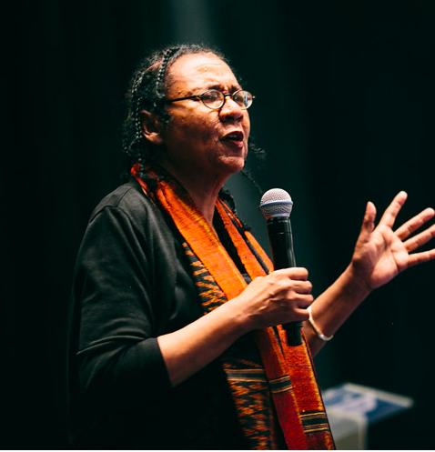 A woman with  cornrows wearing glasses and an orange scarf over dark clothes holds a microphone while gesturing towards a crowd 
