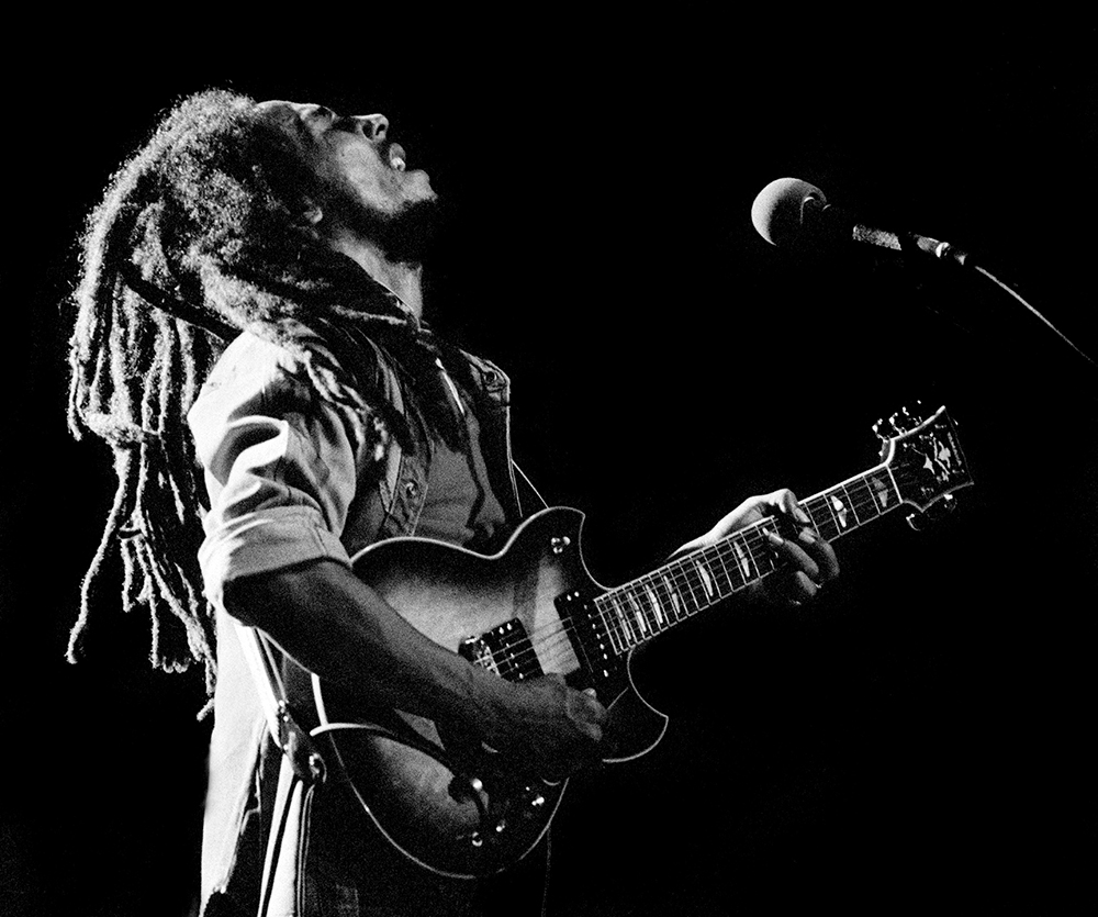 Bob Marley sings while standing and playing guitar in front of a microphone