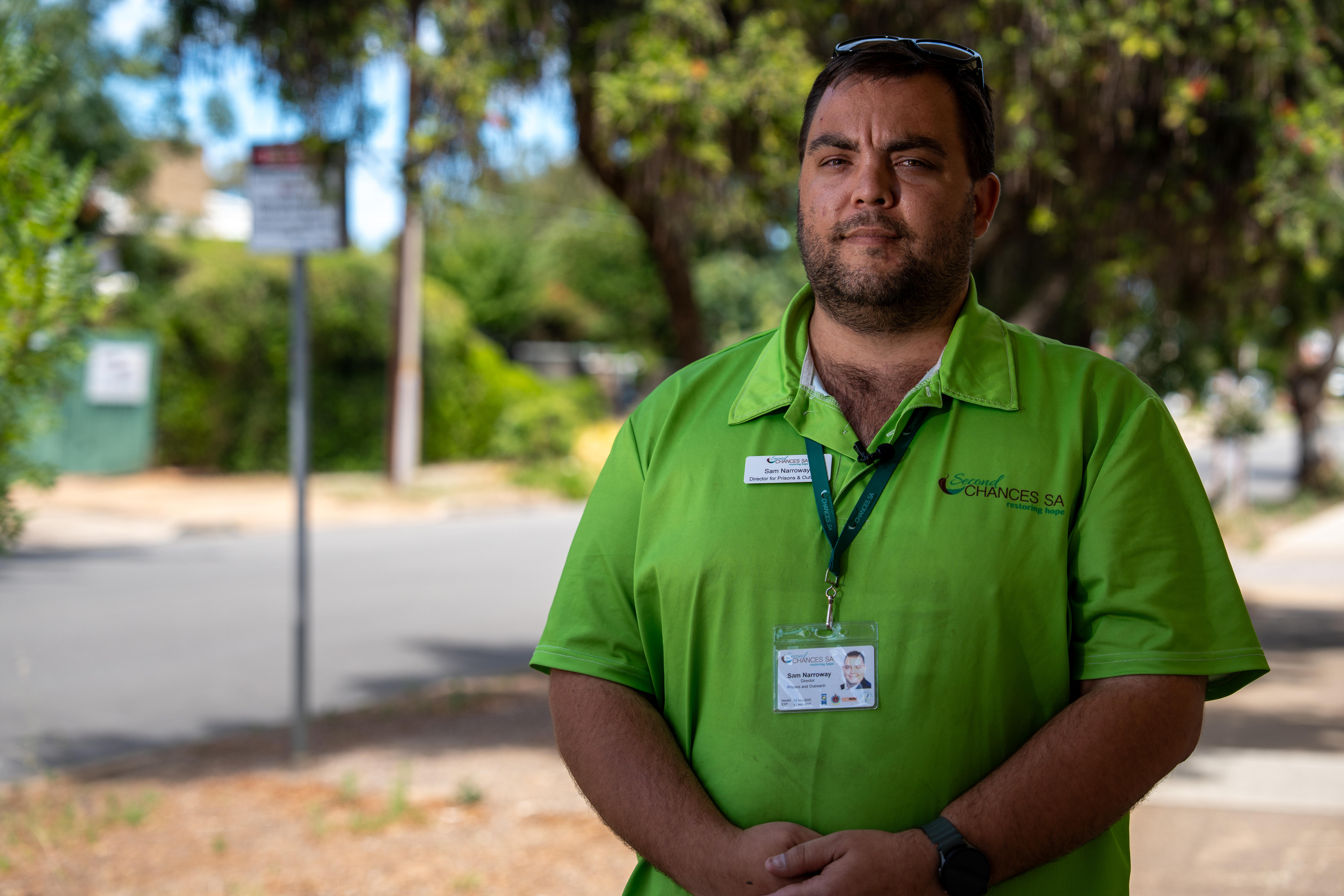 Un hombre con un polo verde con una etiqueta con su nombre en el frente se encuentra en una calle arbolada.