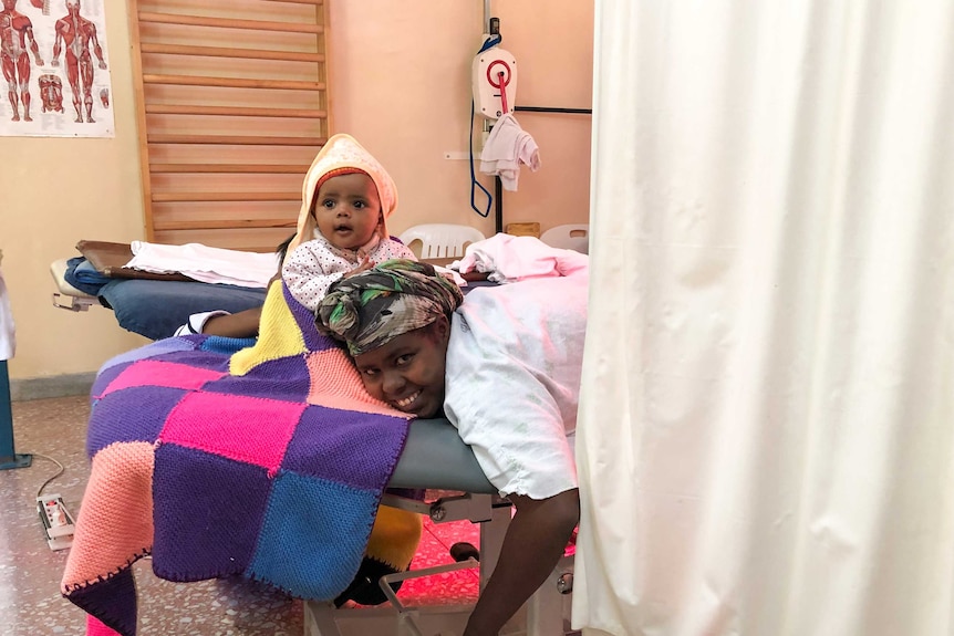 A woman laying on an examination table with a baby sitting next to her.