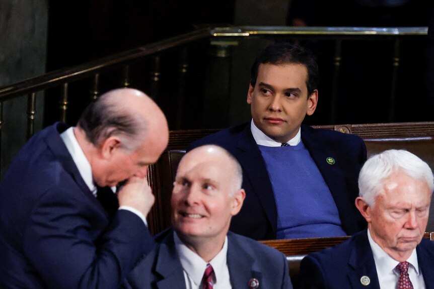 George Santos wearing a jumper and suit sits in Congress looking over as two men laugh and whisper