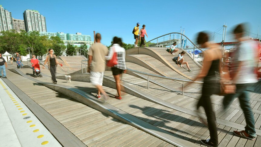 People walk on a boardwalk that has small hills, or waves. Multistorey buildings and bridge in background.