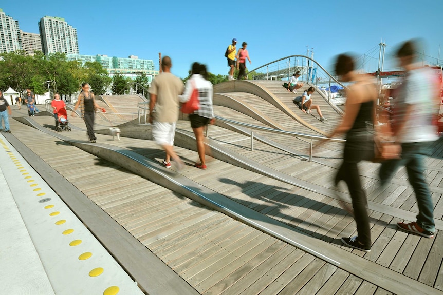 People walk on a boardwalk that has small hills, or waves. Multistorey buildings and bridge in background.