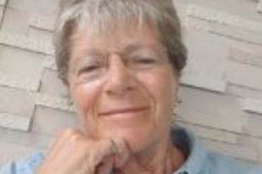 A woman with grey hair and glasses wearing a blue shirt, rests her head on one hand and smiles.