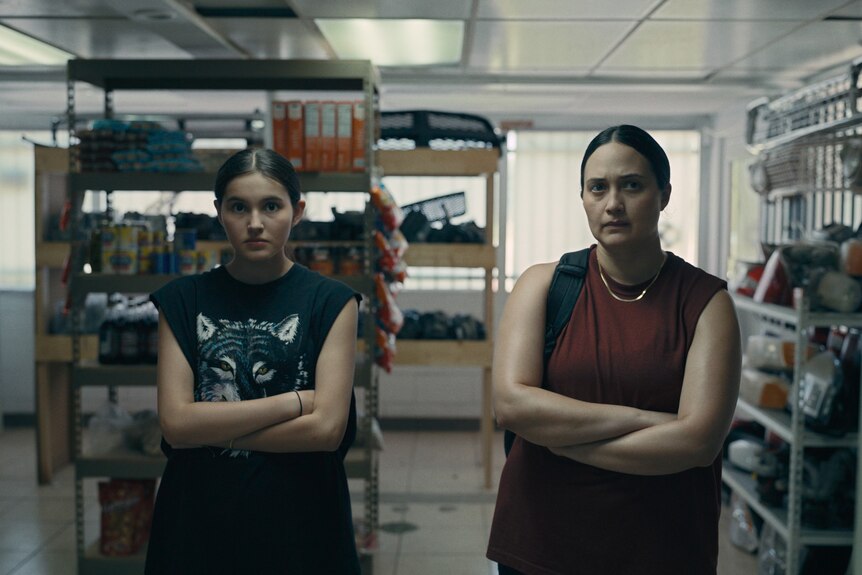 A woman and a teenage girl both stand with their arms crossed, looking serious, inside a convenience store.