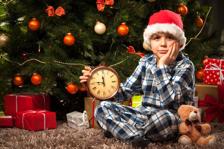 Một cậu bé cầm đồng hồ và ngồi trước cây thông Noel trông chán nản khi chờ đợi thời gian mở cửa hiện tại