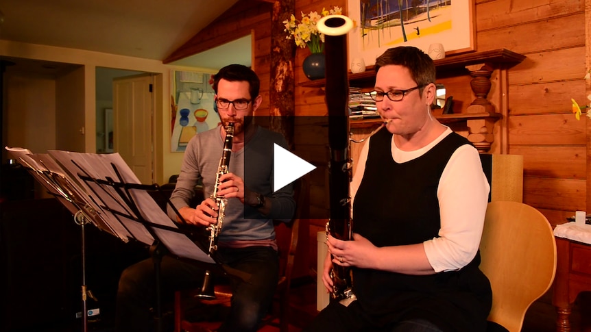 A bassoon player and a clarinet player perform in a living room with daffodils in the background.