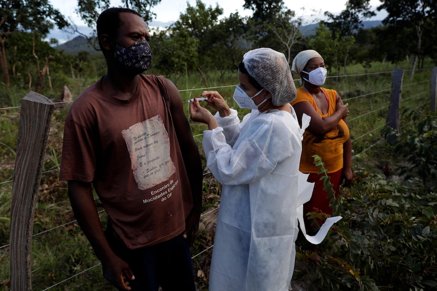 A man stands as he receives a vaccine by a nurse in PPE on a rural property