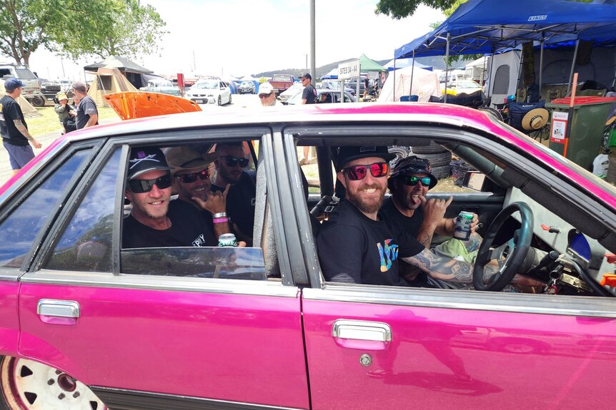 Three men in a bright pink 1986 Holden Commodore car with windows down, one is doing the shaka hand gesture.