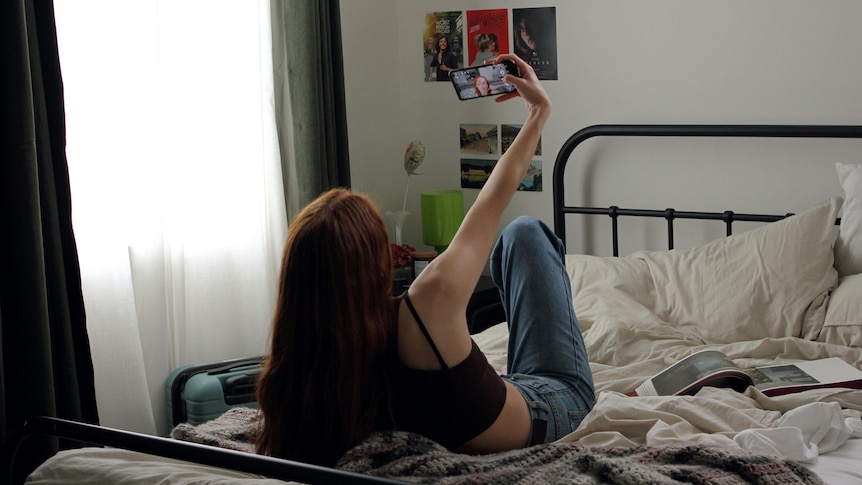 A woman sits on her bed, taking a 'selfie' on her phone as she talks to someone.