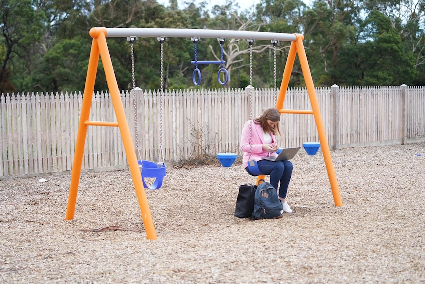 Woman sitting on a swing with laptop on her knees, looking at mobile phone.