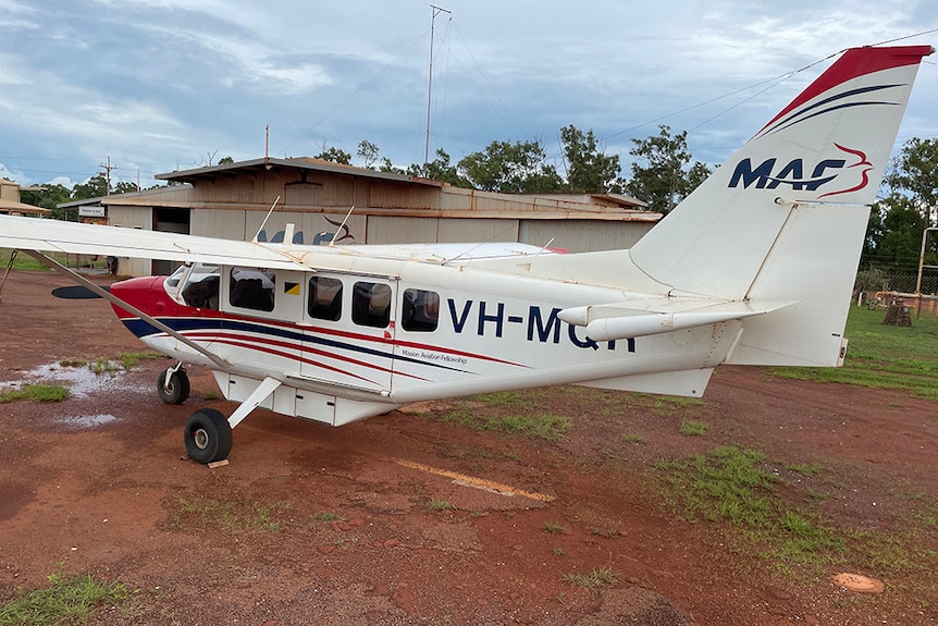 Small white plane sits stationary on red dirt in front of small building