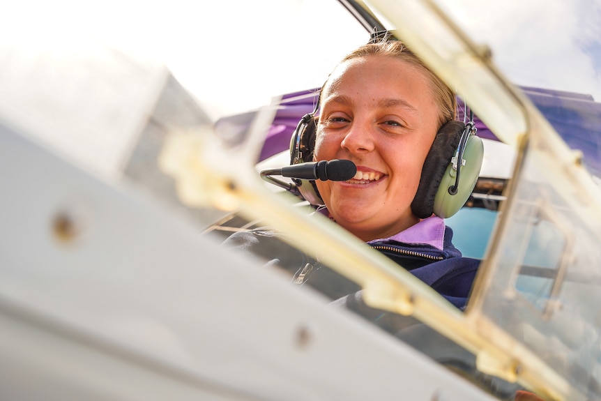 A girl sits in an open cockpit on a sunny day with a headset on grinning.