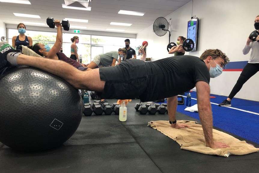 A man with his legs stretched across an exercise ball at a gym wearing a mask.