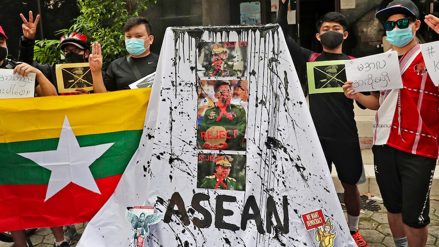 Protesters stand behind a white sheet emblazoned with Myanmar's military leaders and the word 'ASEAN', dashed with black paint.