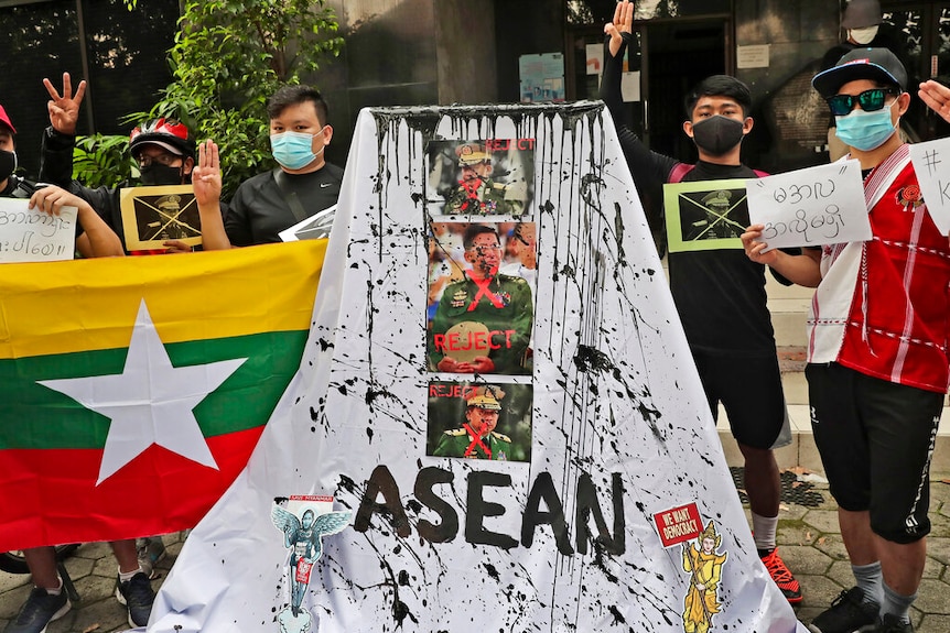 Protesters stand behind a white sheet emblazoned with Myanmar's military leaders and the word 'ASEAN', dashed with black paint.