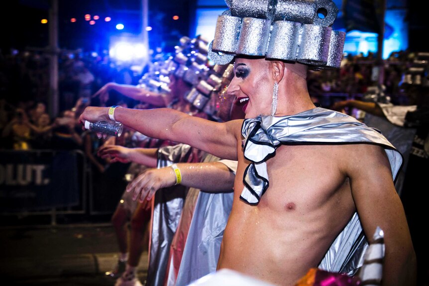 Shirtless men in silver hats and capes, covered in glitter, dance in the parade.