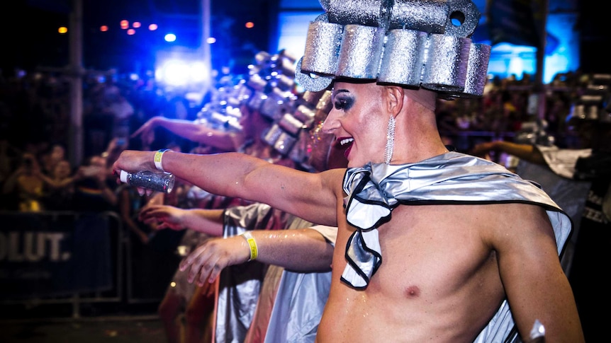 Shirtless men in silver hats and capes, covered in glitter, dance in the parade.