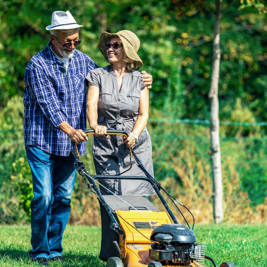 An older couple wearing wide-brimmed hats mow the lawn together joyfully on a bright day.