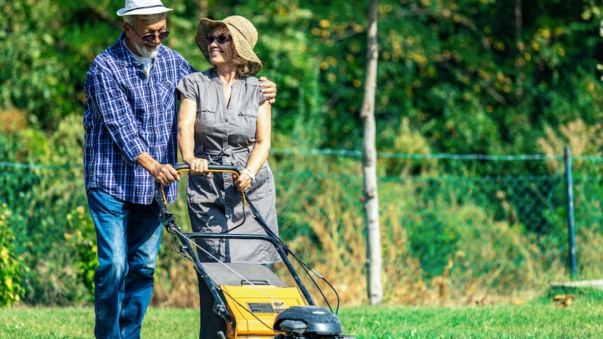 An older couple wearing wide-brimmed hats mow the lawn together joyfully on a bright day.