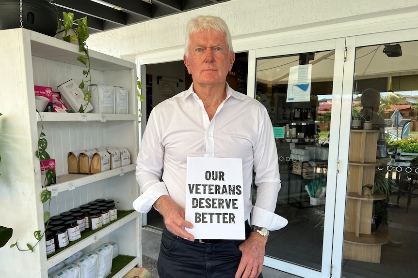 A grey-haired serious man standing with a sign which reads: 'our veterans deserve better'.