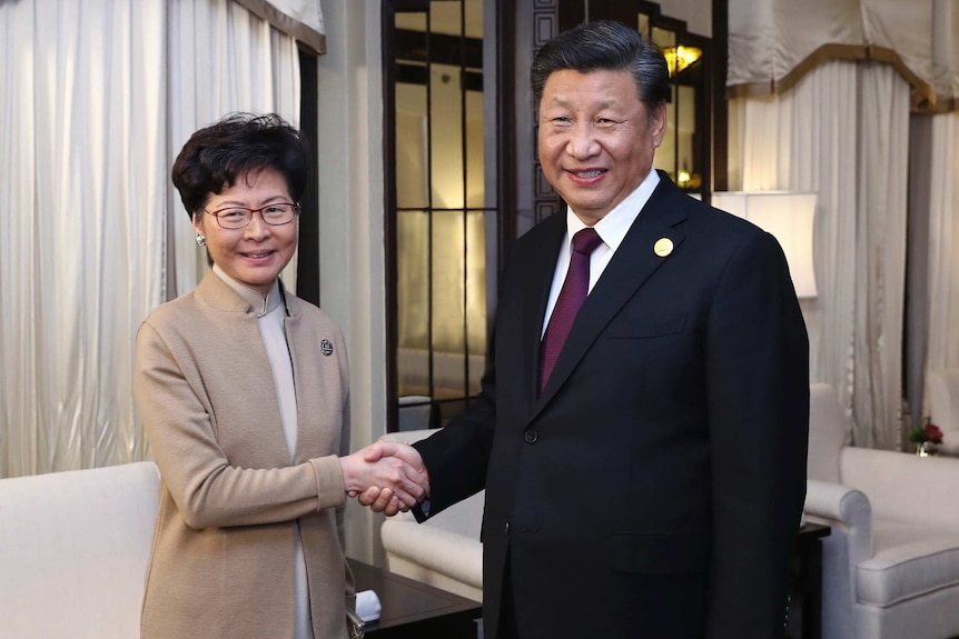 Carrie Lam po lewej w beżowej sukience i kurtce, a Xi Jinping po prawej w czarnym garniturze, oboje uśmiechają się do kamery, ściskając ręce