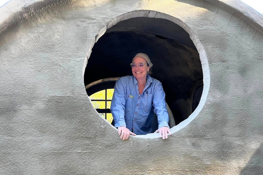 A middle-aged woman with glasses and a blue shirt stands inside a dome-shaped house, looking through a round window and smiling