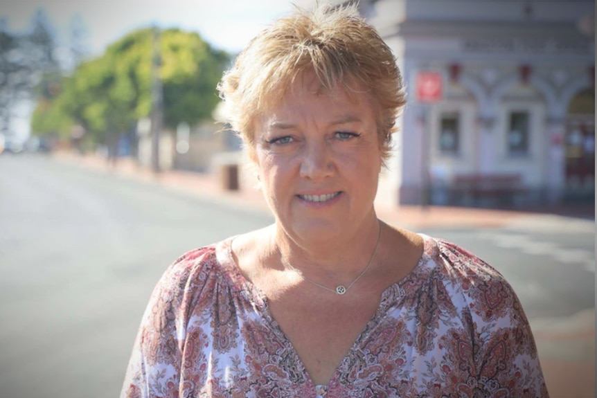 A woman with short blonde hair smiles at the camera with a street blurred behind her