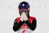 A woman wearing Olympic ski gear, including a helmet, holds her hands in front of her nose and mouth.