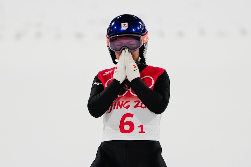 A woman wearing Olympic ski gear including a helmet holds her hands in front of her nose and mouth.