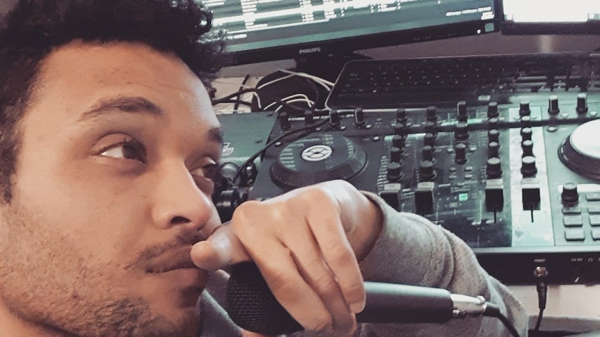 Man sits in front of computer and mixing board with microphone in left hand.