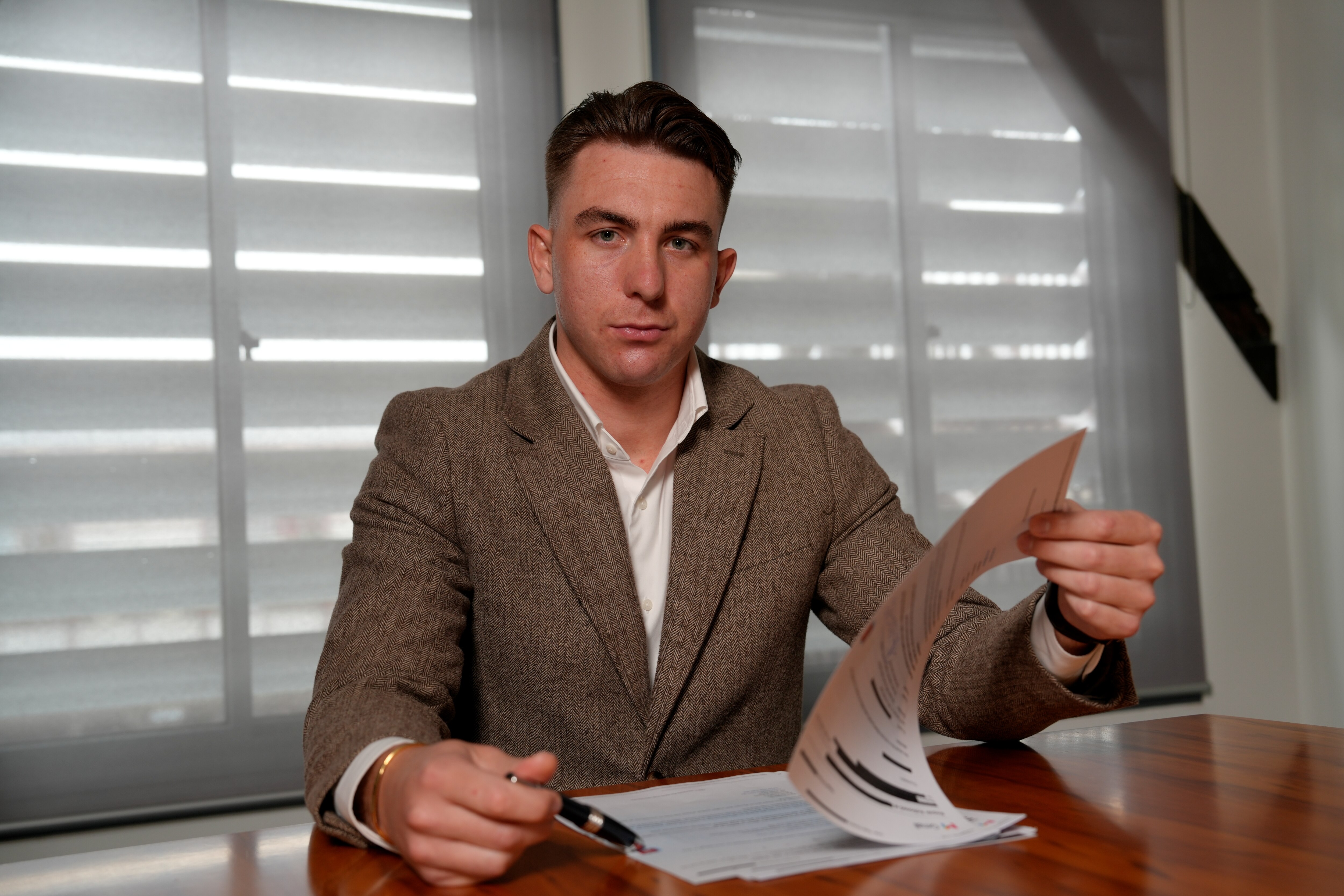 A man in his late 20s wearing a tweed suit jacket and white business shirt sits at a wooden table reviewing documents.