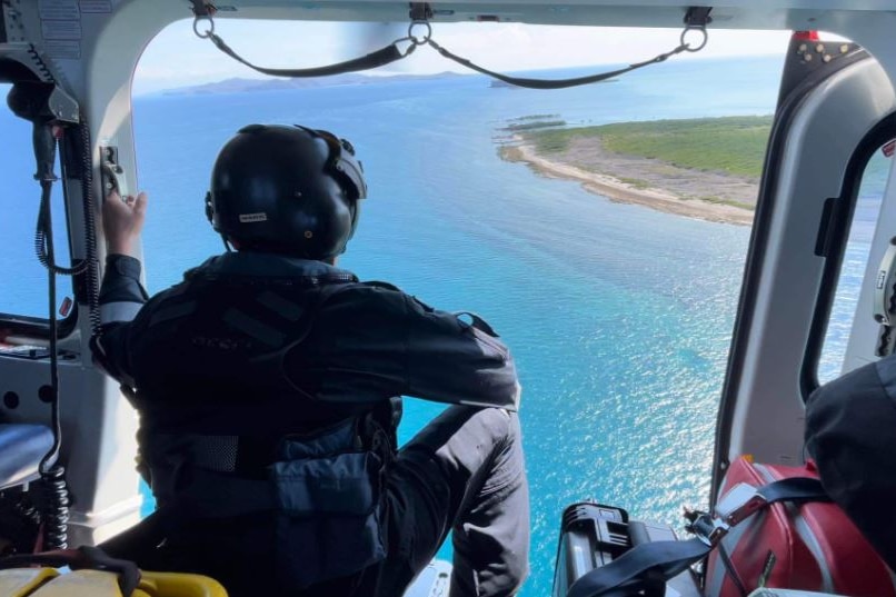 A person sitting in the open doorway of a helicopter looking over the ocean and the edge of an island.
