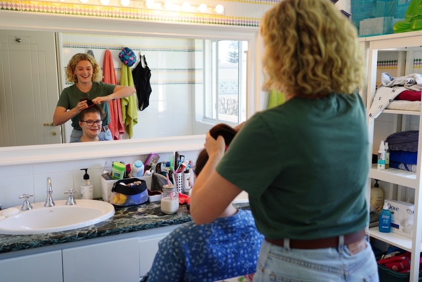 Two women in a bathroom smiling and hair styling in the mirror