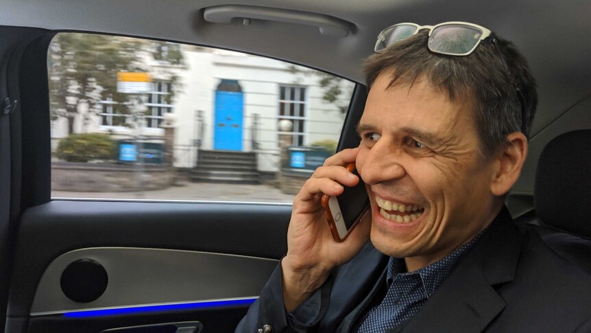 Didier Queloz, wearing a suit, sits in the back of a car while speaking into his mobile. He has a huge grin one his face.