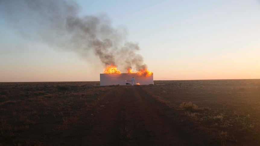 A man dressed in white sets a white wall, in the middle of a red dirt road, on fire.