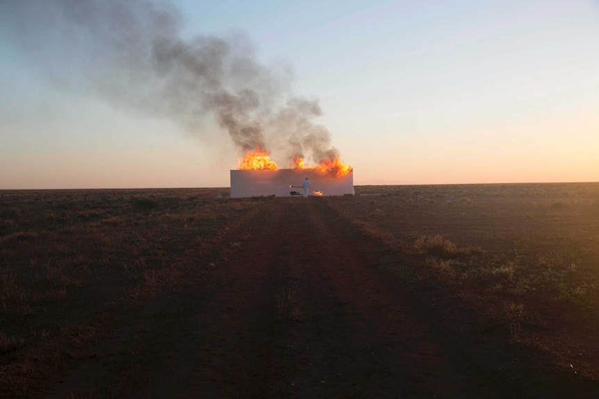 A man dressed in white sets a white wall, in the middle of a red dirt road, on fire.