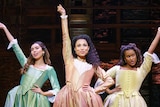 Stage shot showing three women in pastel-coloured 18th century gowns, each with one hand on hip and the other in the air.