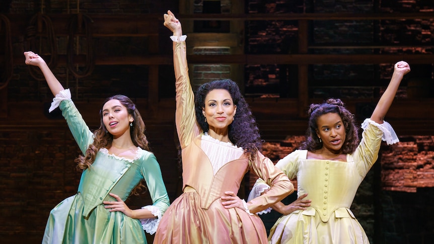 Stage shot showing three women in pastel-coloured 18th century gowns, each with one hand on hip and the other in the air.